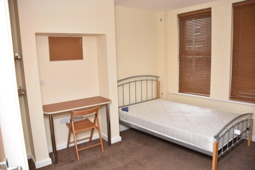 A simple bedroom featuring a metal bed frame with a bare mattress, a wooden desk, and a chair, beside window blinds.