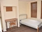 A simple bedroom featuring a metal bed frame with a bare mattress, a wooden desk, and a chair, beside window blinds.