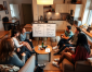 A group discussion takes place in a cozy living room, with a whiteboard listing pros and cons. Drinks and snacks are on the table.