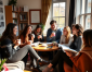 A group of six people enjoy drinks and converse around a coffee table, with books and a small plant in a sunlit, cozy room.